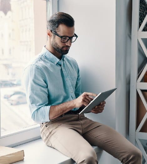 A man in glasses sitting on a window sill, engrossed in using a tablet device.